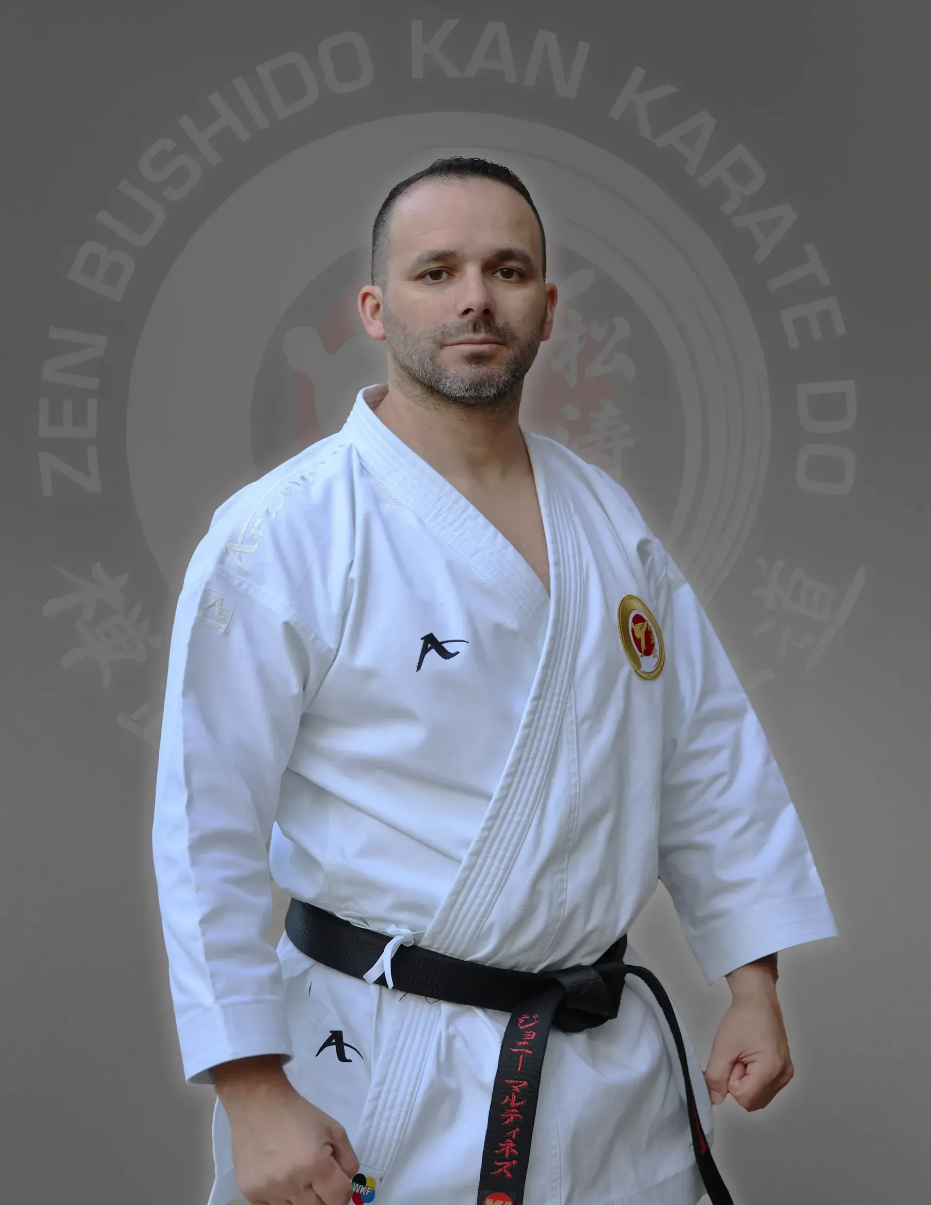 Man in karate uniform, black belt, standing in front of a dojo logo.