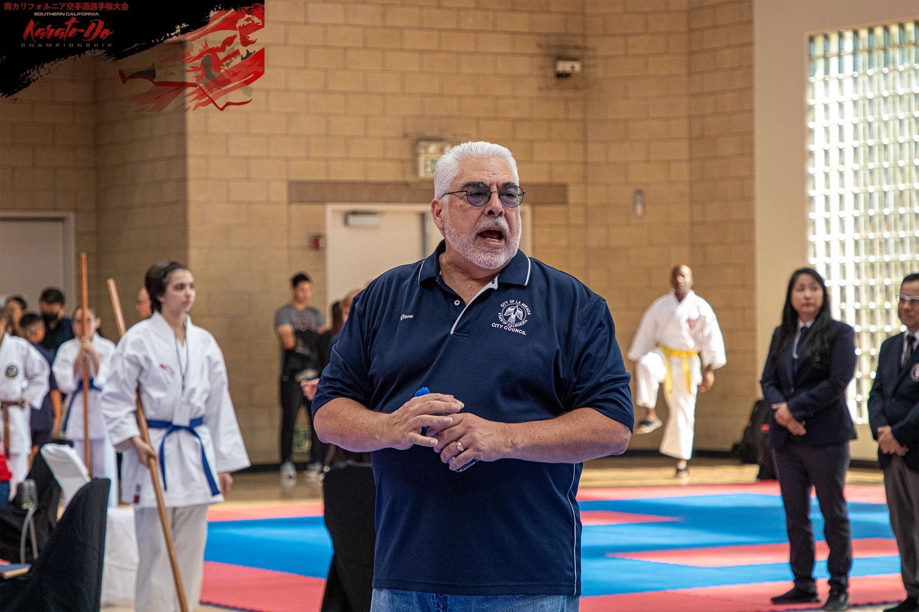 A man is standing on a karate mat talking to a group of people.