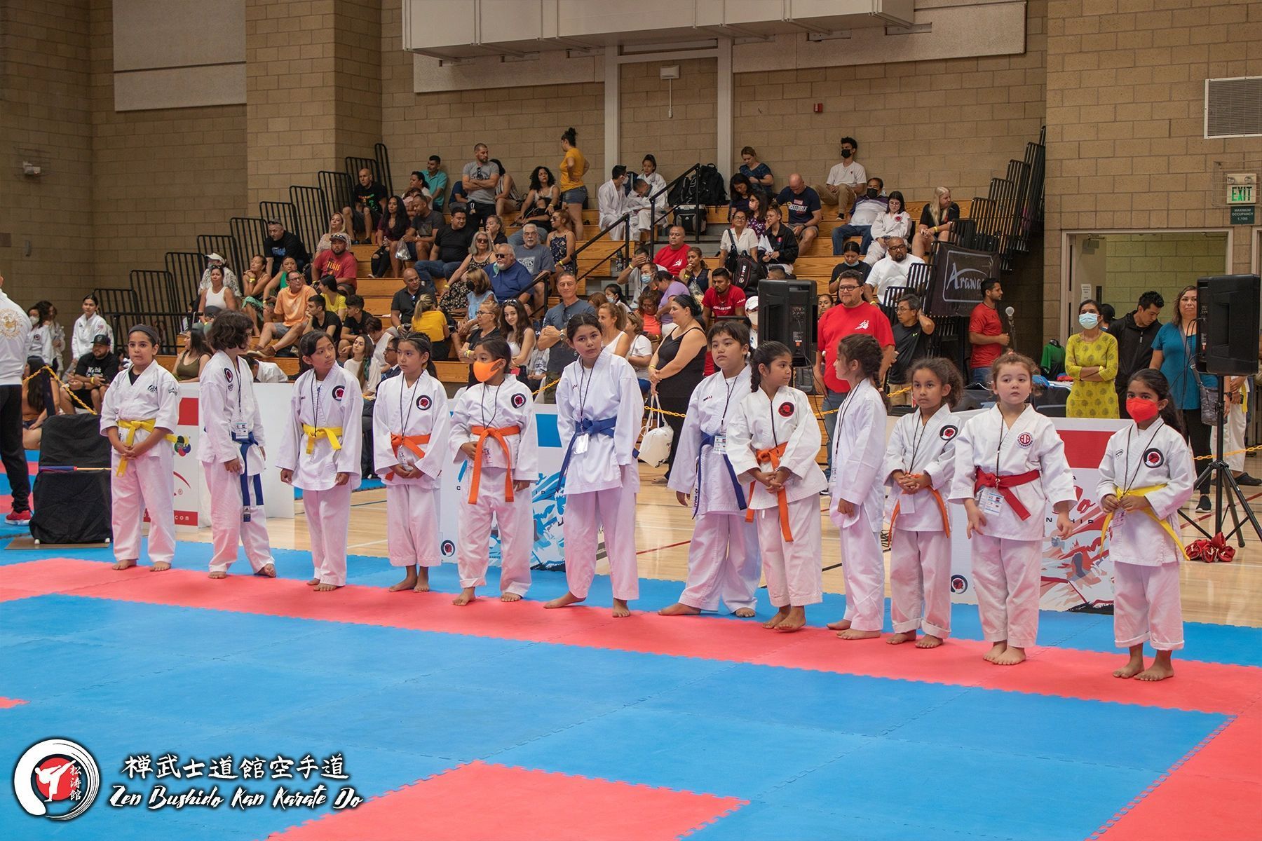 A group of children are standing on a karate mat in front of a crowd.