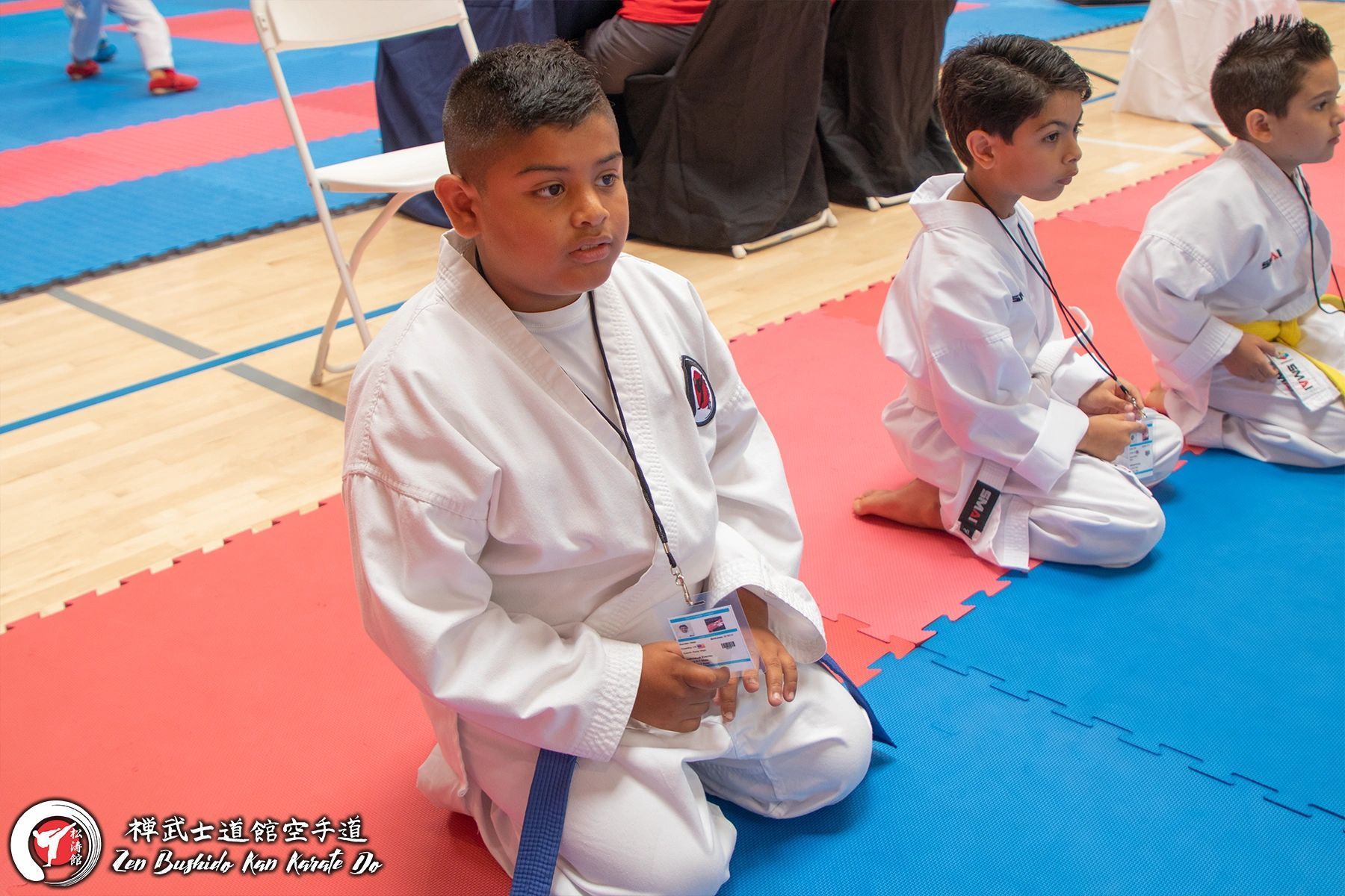 Three young boys in karate uniforms are kneeling on a mat in a gym.