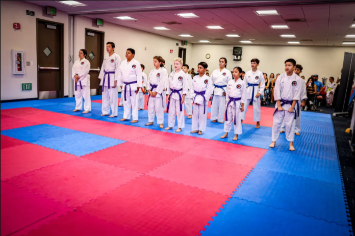 A group of people are standing on a karate mat in a gym.