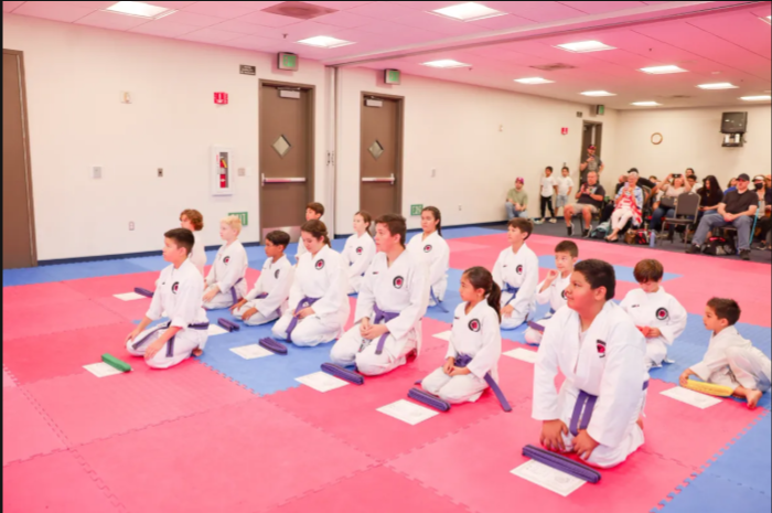 A group of children are sitting on a karate mat in a gym.