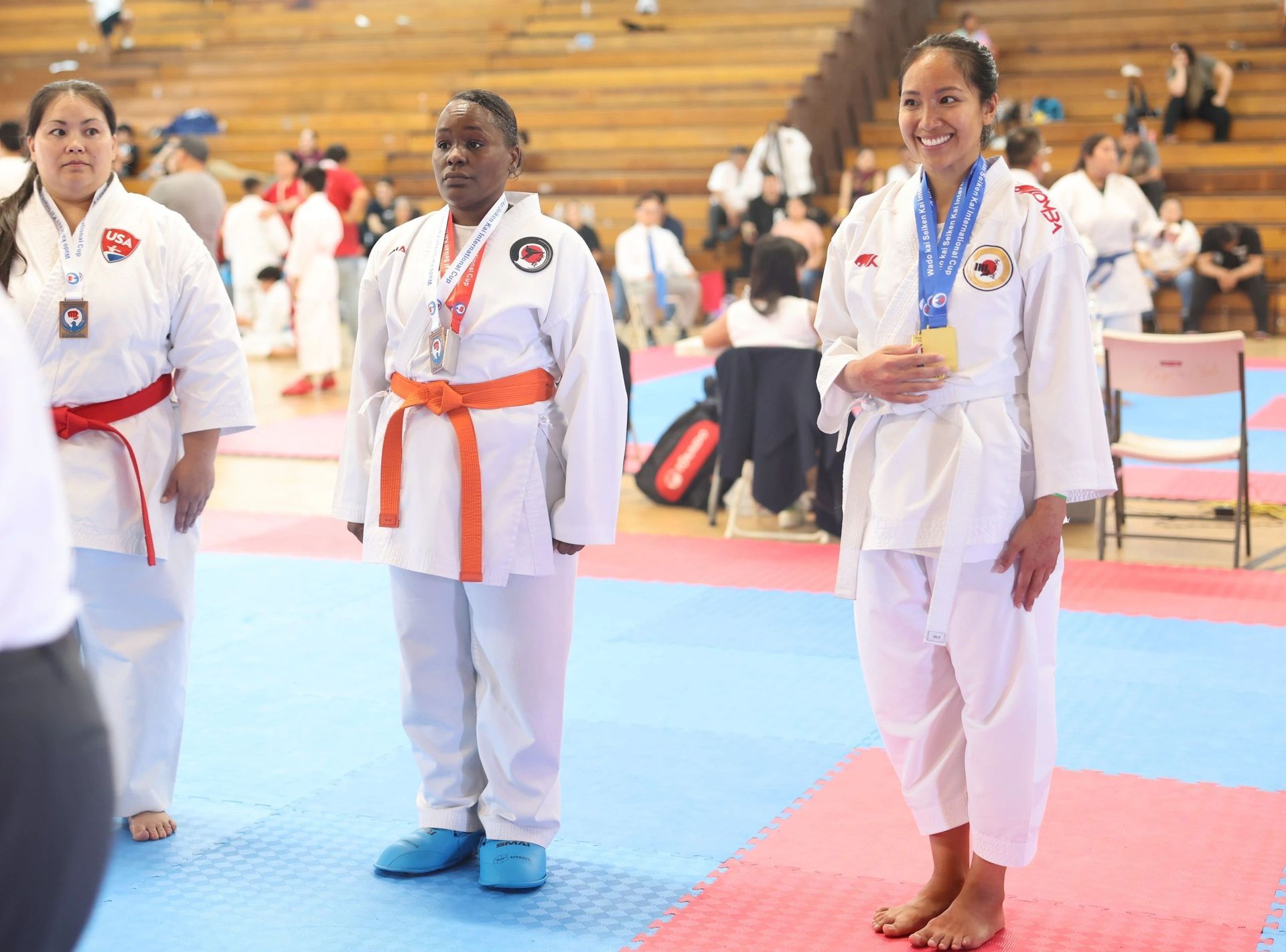 Three women in karate uniforms are standing on a mat in a gym.