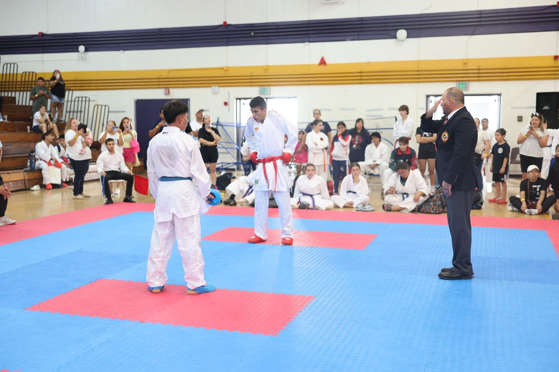 A group of people are standing on a karate mat in a gym.