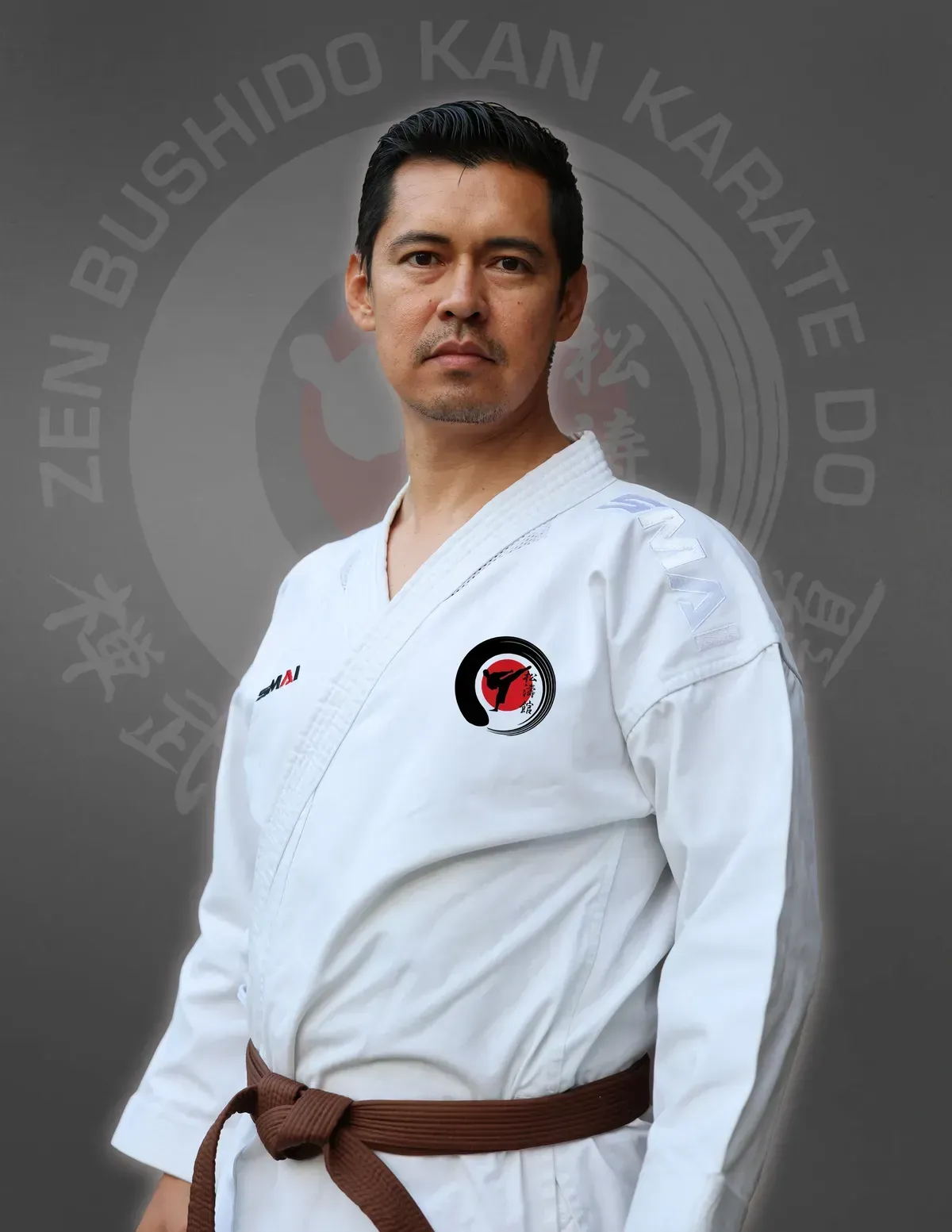 Man in karate uniform with brown belt, standing in front of a dojo emblem.