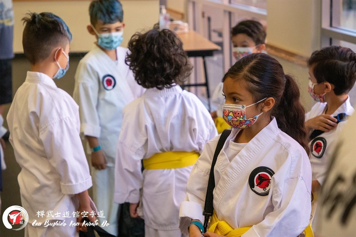 A group of children wearing masks and karate uniforms are standing in a room.