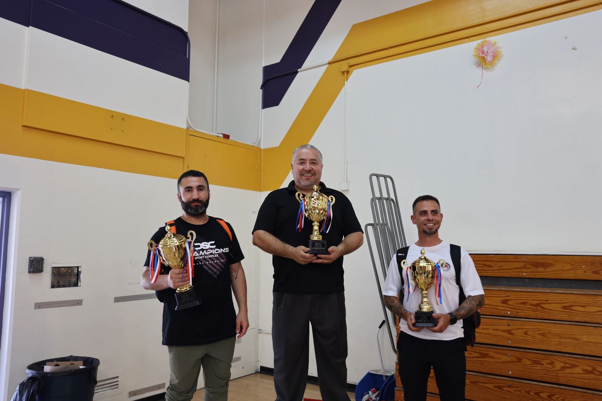 Three men are standing in a gym holding trophies.