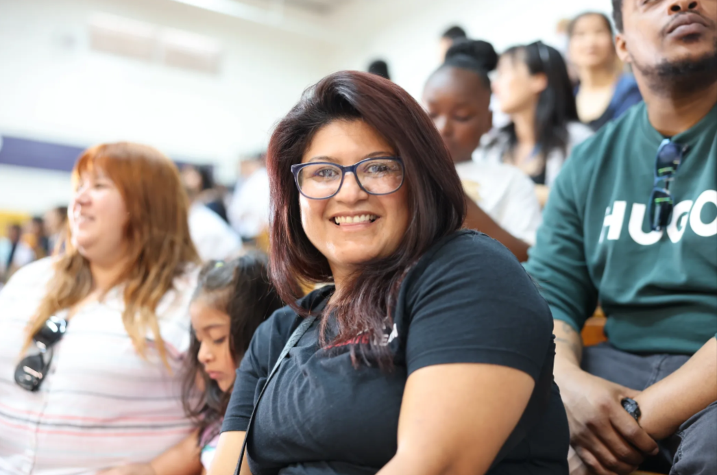 A woman wearing glasses is smiling while sitting in a crowd of people.