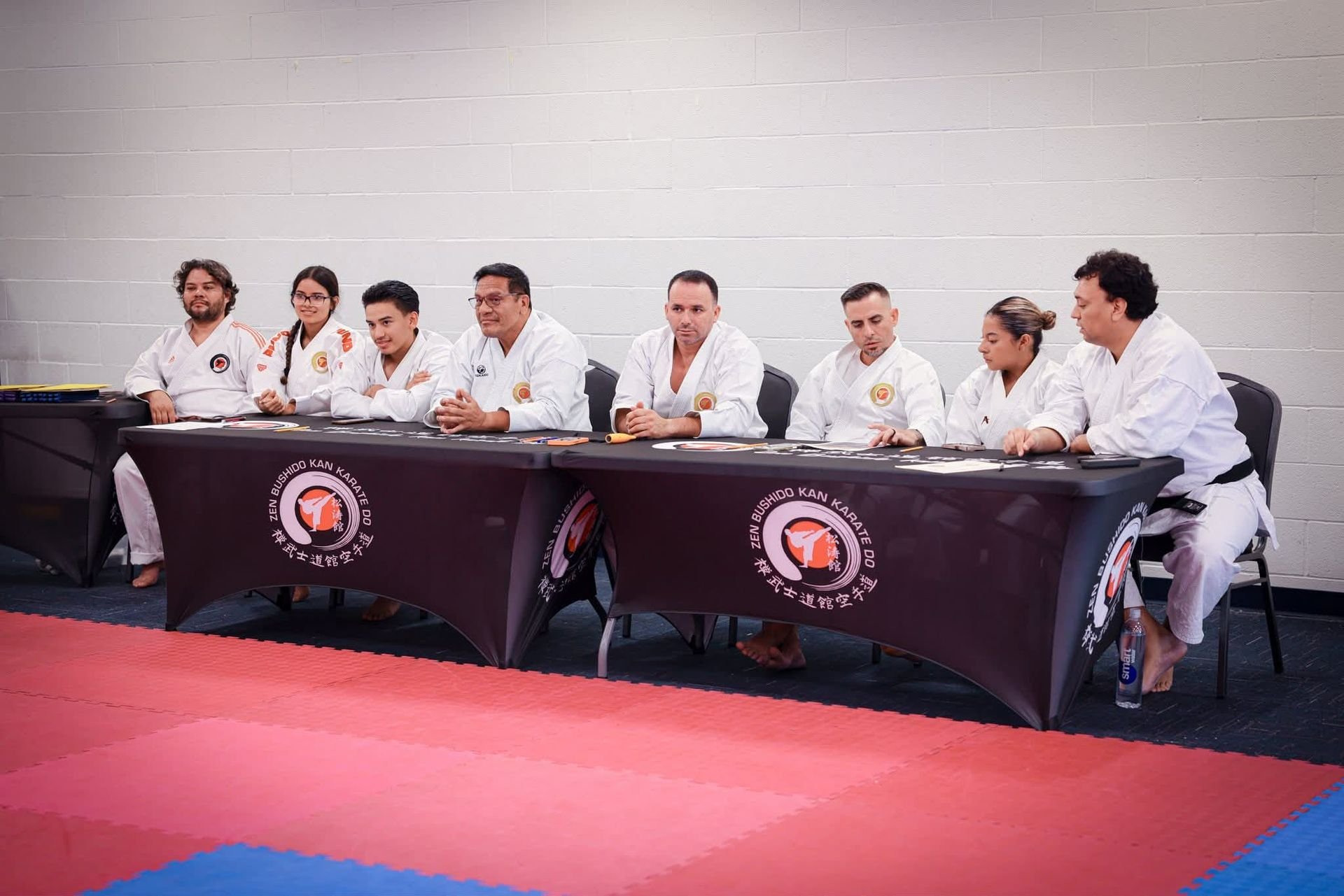 Karate judges at a table, wearing white uniforms, assessing a competition on a red mat.