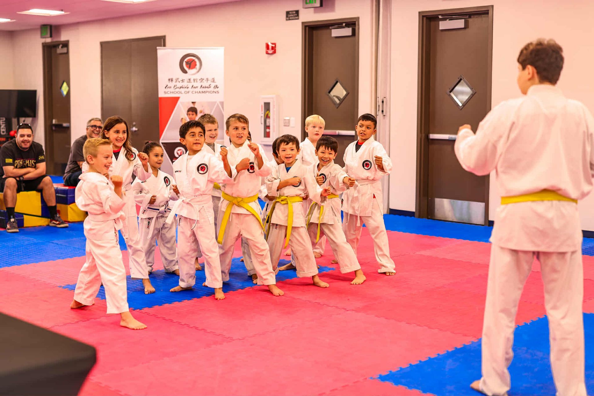 Children in karate uniforms practice in a gym, following an instructor.
