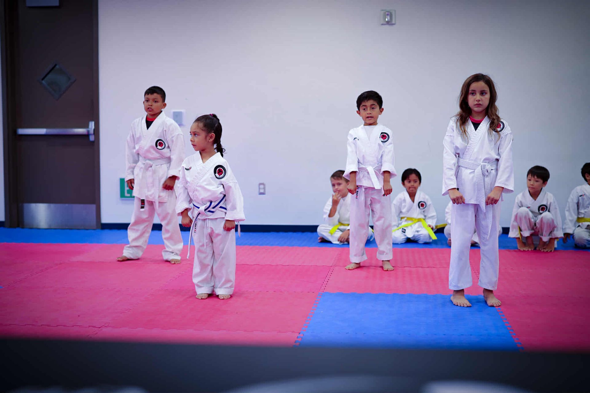 Children in white karate uniforms stand on a blue and red mat in a dojo, practicing.