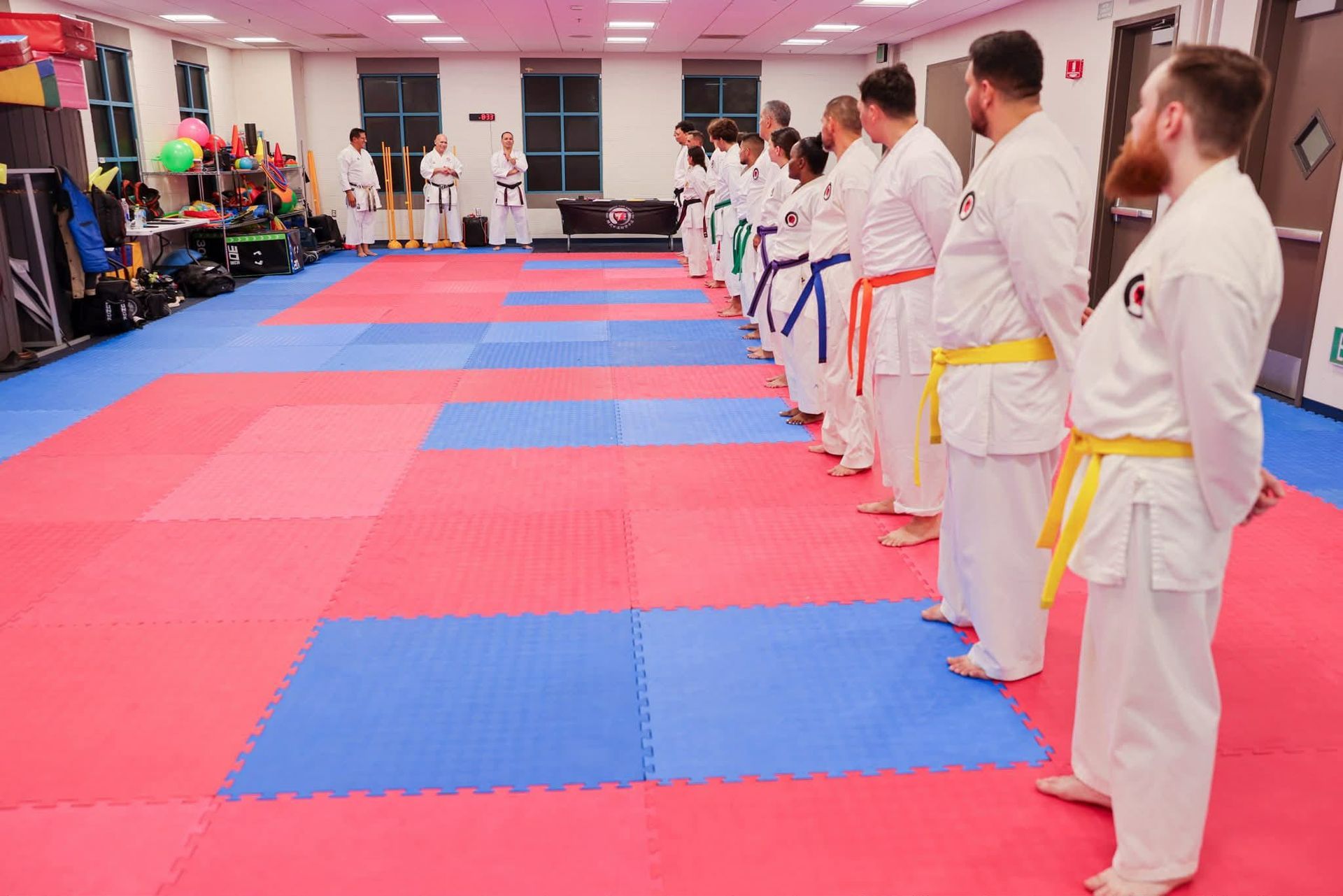 Karate practitioners in white uniforms, lined up on a blue and red mat, facing away from the camera.