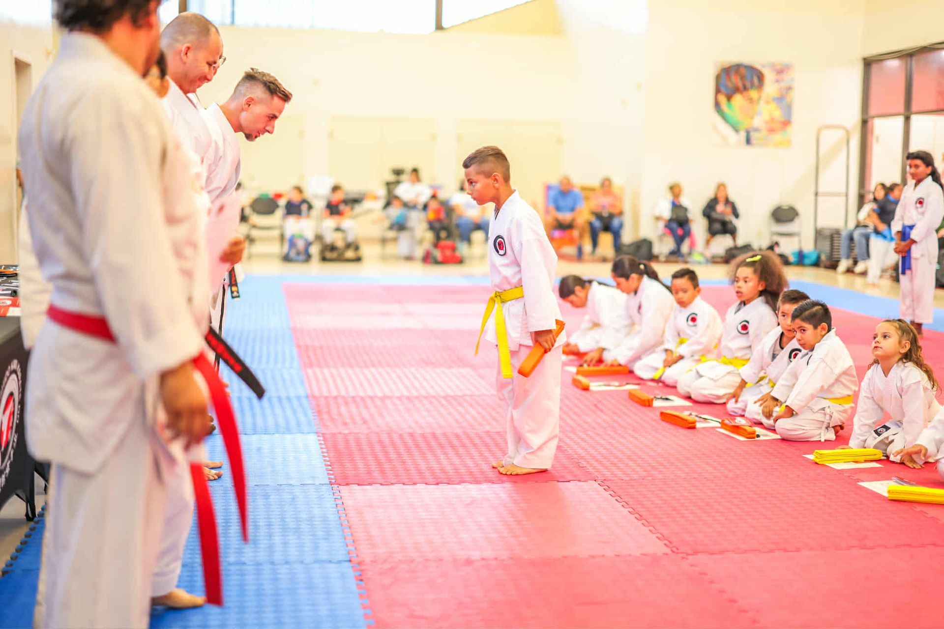 Karate students bowing at a dojo; yellow belt, red belt, and spectators in background.