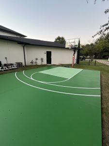 A green basketball court with a basketball hoop in front of a house.