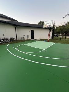 A green basketball court with a white hoop in front of a white building.