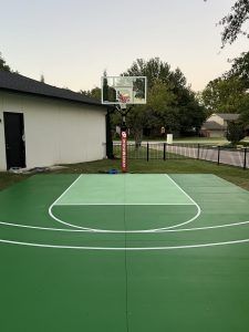 A green basketball court with a basketball hoop in the backyard of a house.