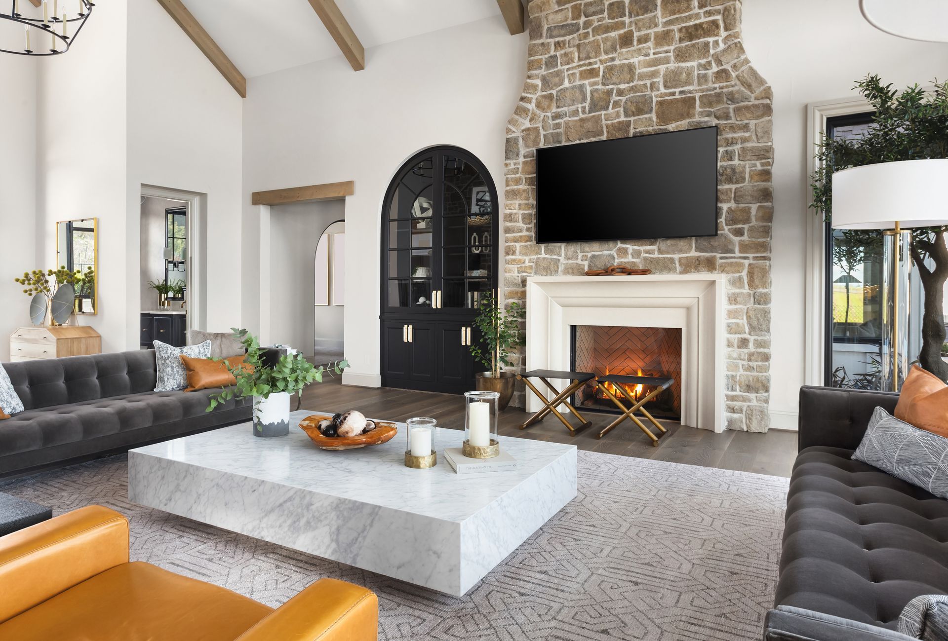 Living room with fireplace, stone wall, TV, and black arched door. Features gray couches, marble coffee table, and wood beams.
