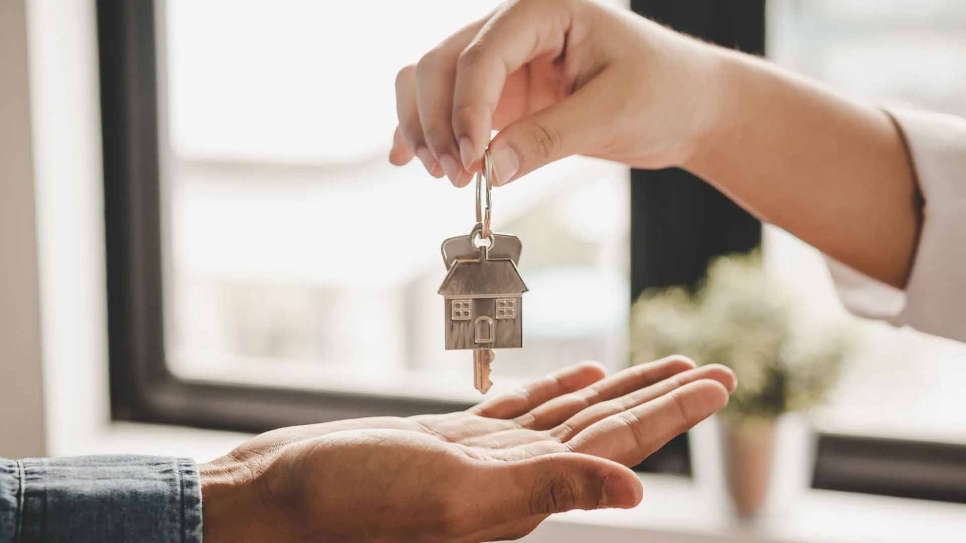 Person handing over a house key with a house-shaped keychain; indoors, bright lighting.