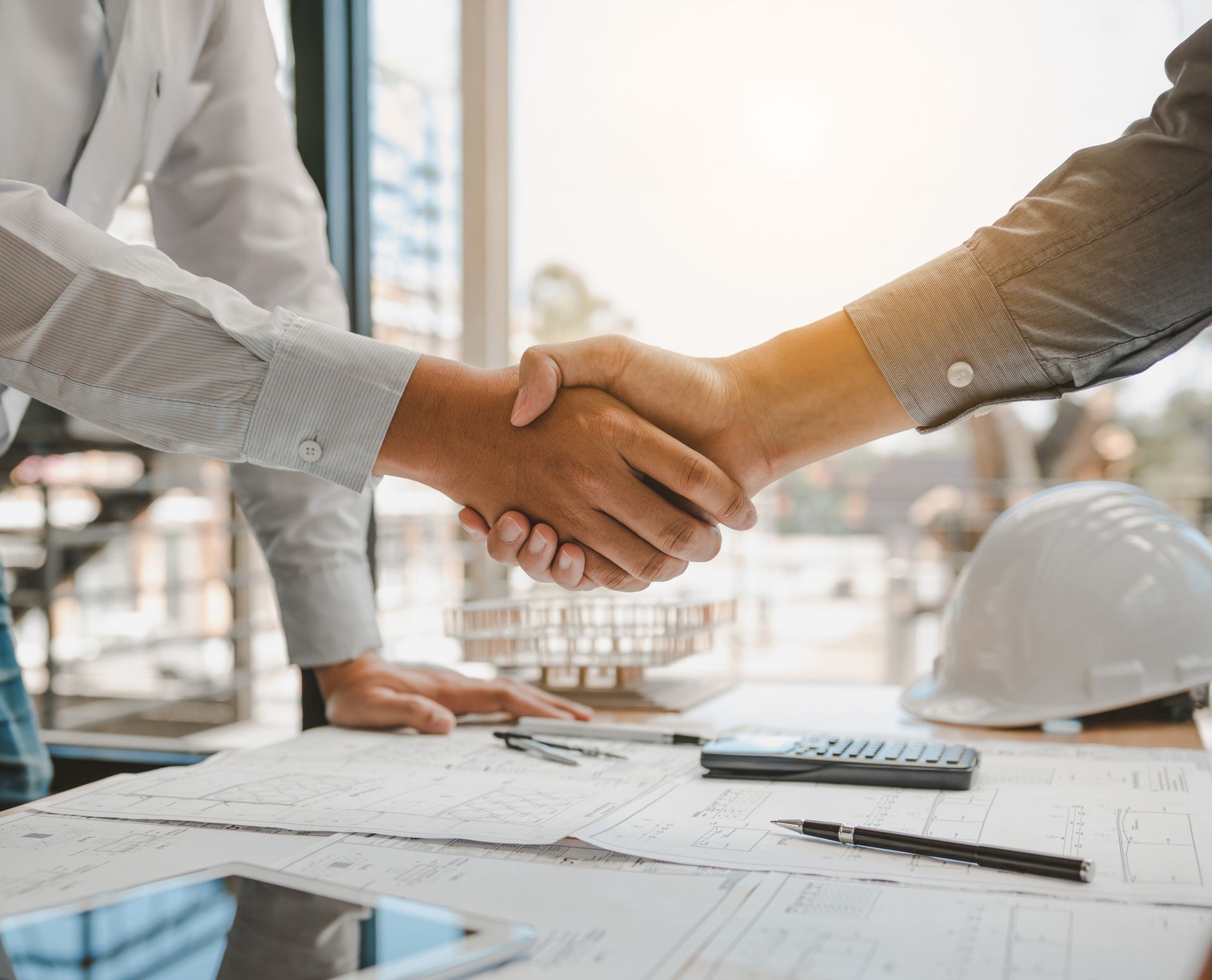 Deux personnes se serrant la main au-dessus de plans et d'un casque de chantier sur un bureau, probablement après un accord.