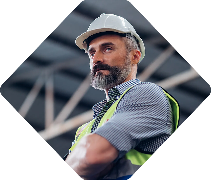 Homme avec une barbe portant un casque et un gilet de sécurité, les bras croisés, l'air pensif dans un entrepôt.