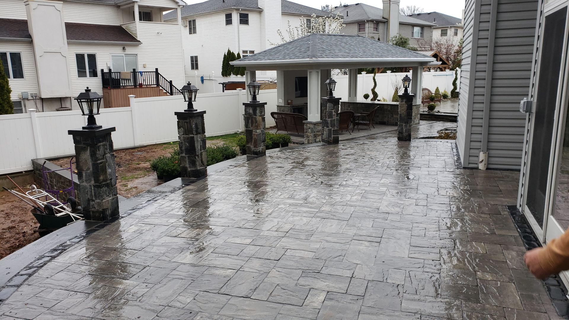 Brick patio with gazebo and lamp posts in a backyard setting; wet ground reflecting the sky.