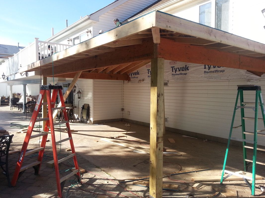 Construction of a wooden patio roof attached to a two-story white house. Ladders and materials visible.