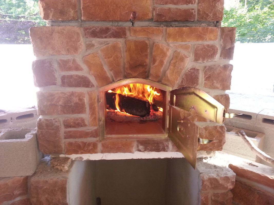 Wood-fired brick oven with an open door and burning logs inside, built with reddish-brown stones.
