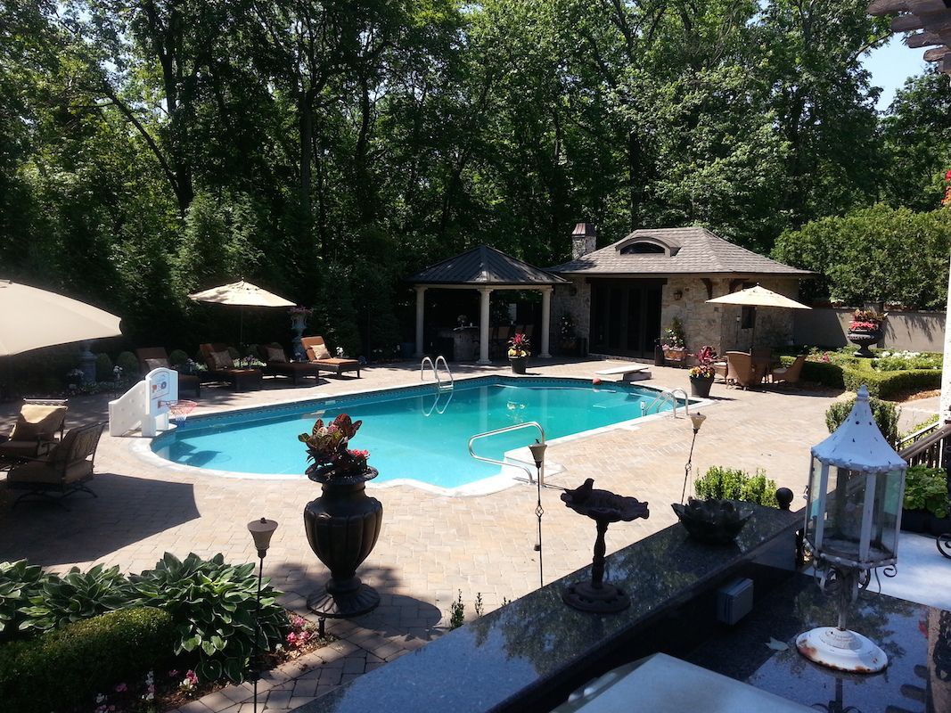 Swimming pool surrounded by stone patio, lush greenery, and a covered pool house.