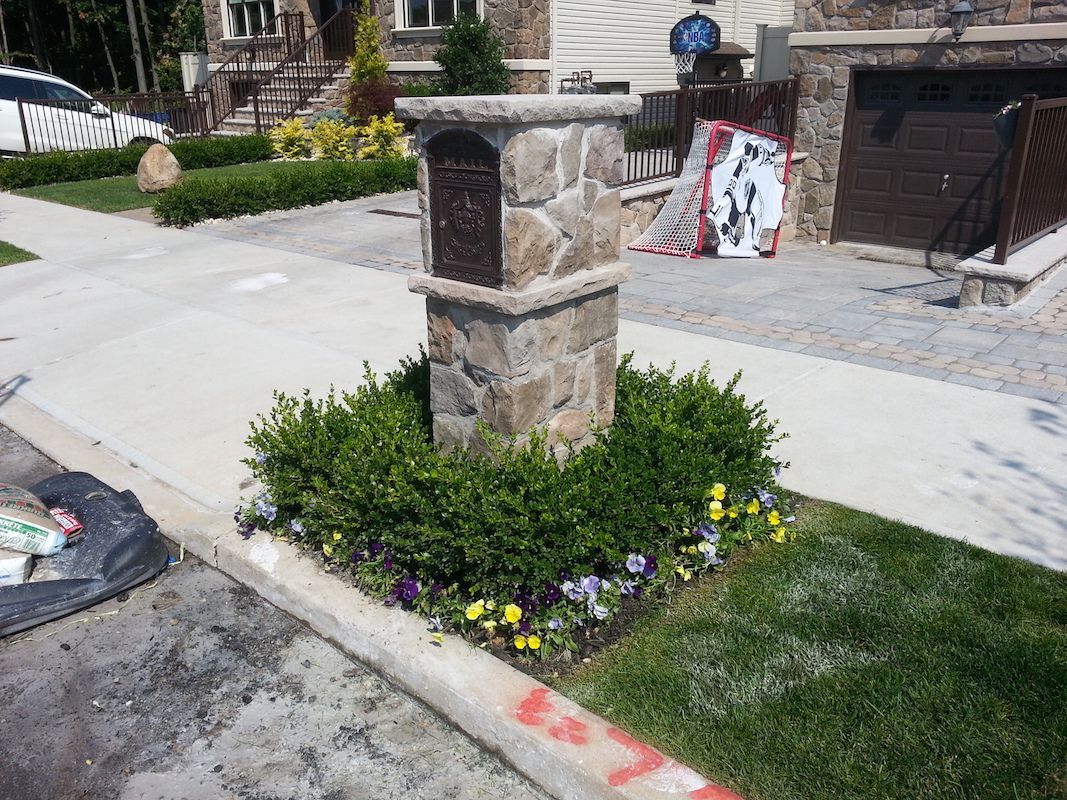 Stone mailbox surrounded by green shrubs and colorful flowers on a concrete sidewalk.