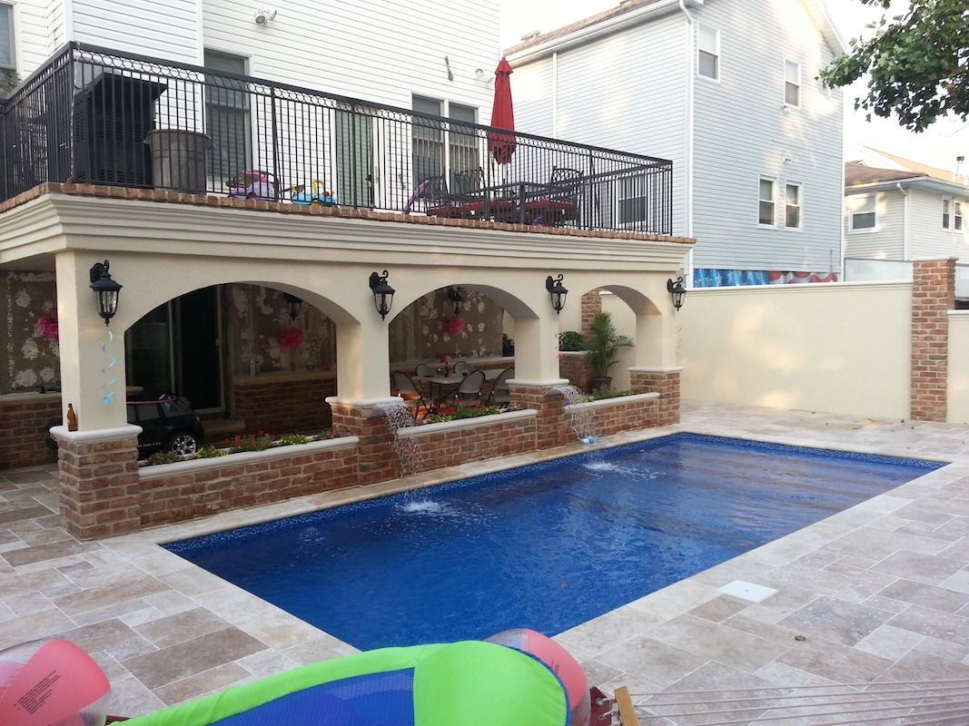 Swimming pool with a patio and a balcony in a backyard. Blue water, tan tiles, red umbrella.