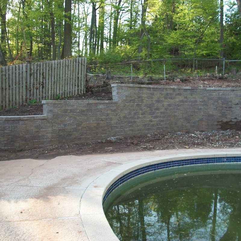 Poolside with retaining walls and a wooden fence, surrounded by trees.