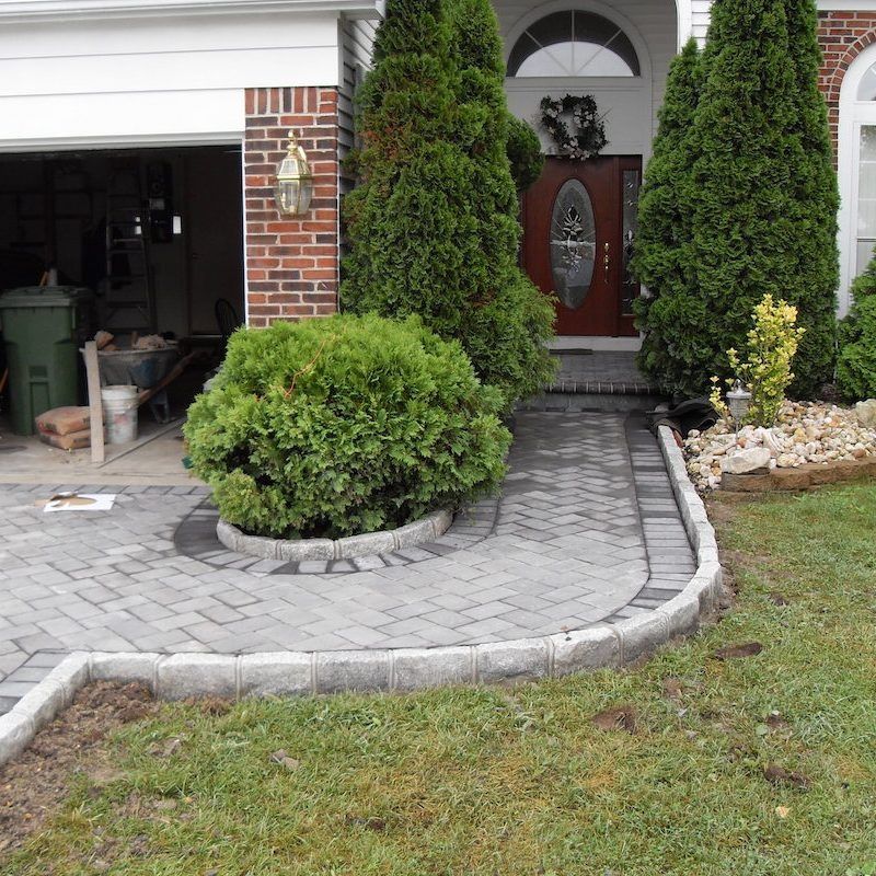 Brick paved pathway leading to a house entrance, with shrubs and green grass in front.