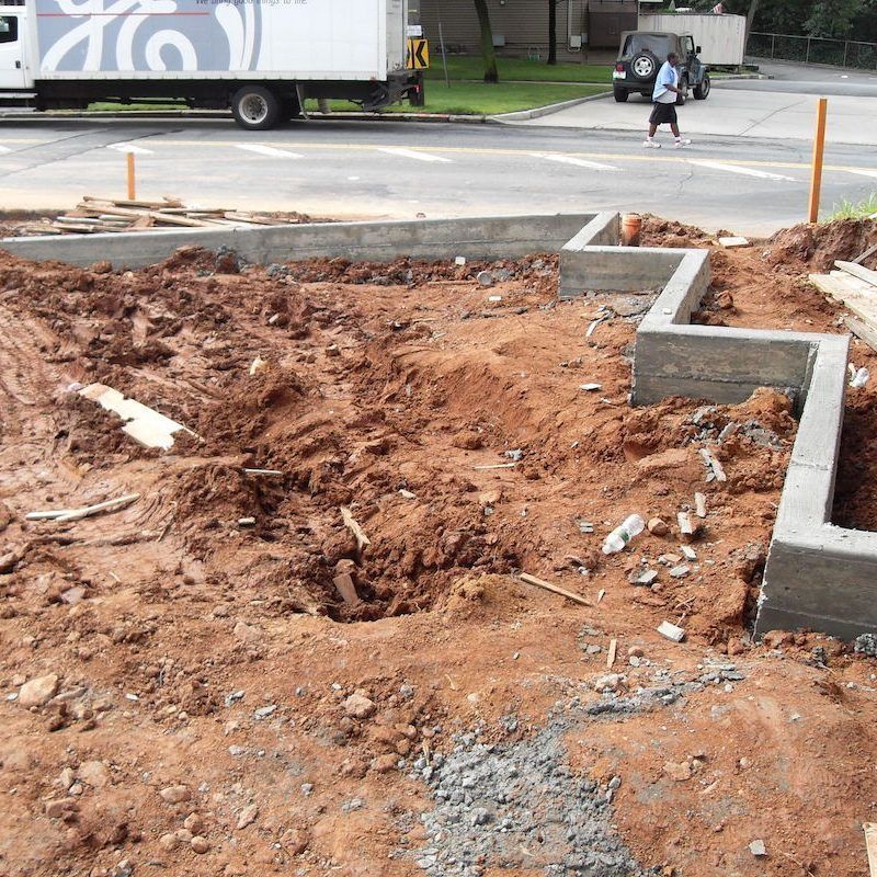 Construction site with concrete foundation and red dirt. A truck, Jeep, and person crossing the street are visible.