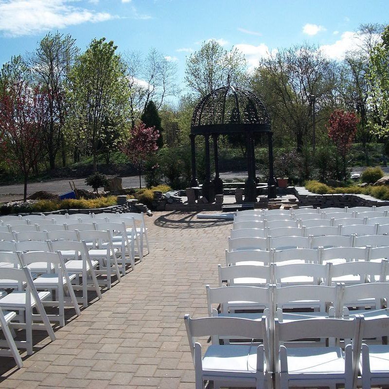 Outdoor wedding ceremony setup with rows of white chairs facing a gazebo on a brick patio.