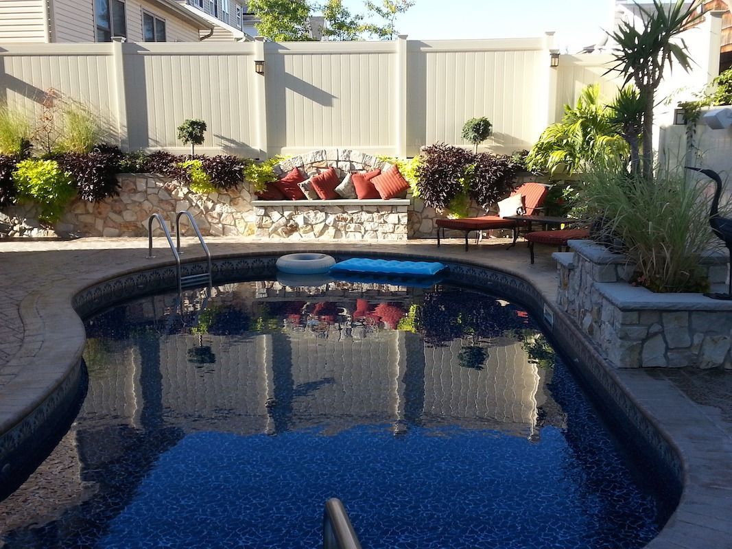Pool with surrounding stone work, beige fence, and lounge area with plants.