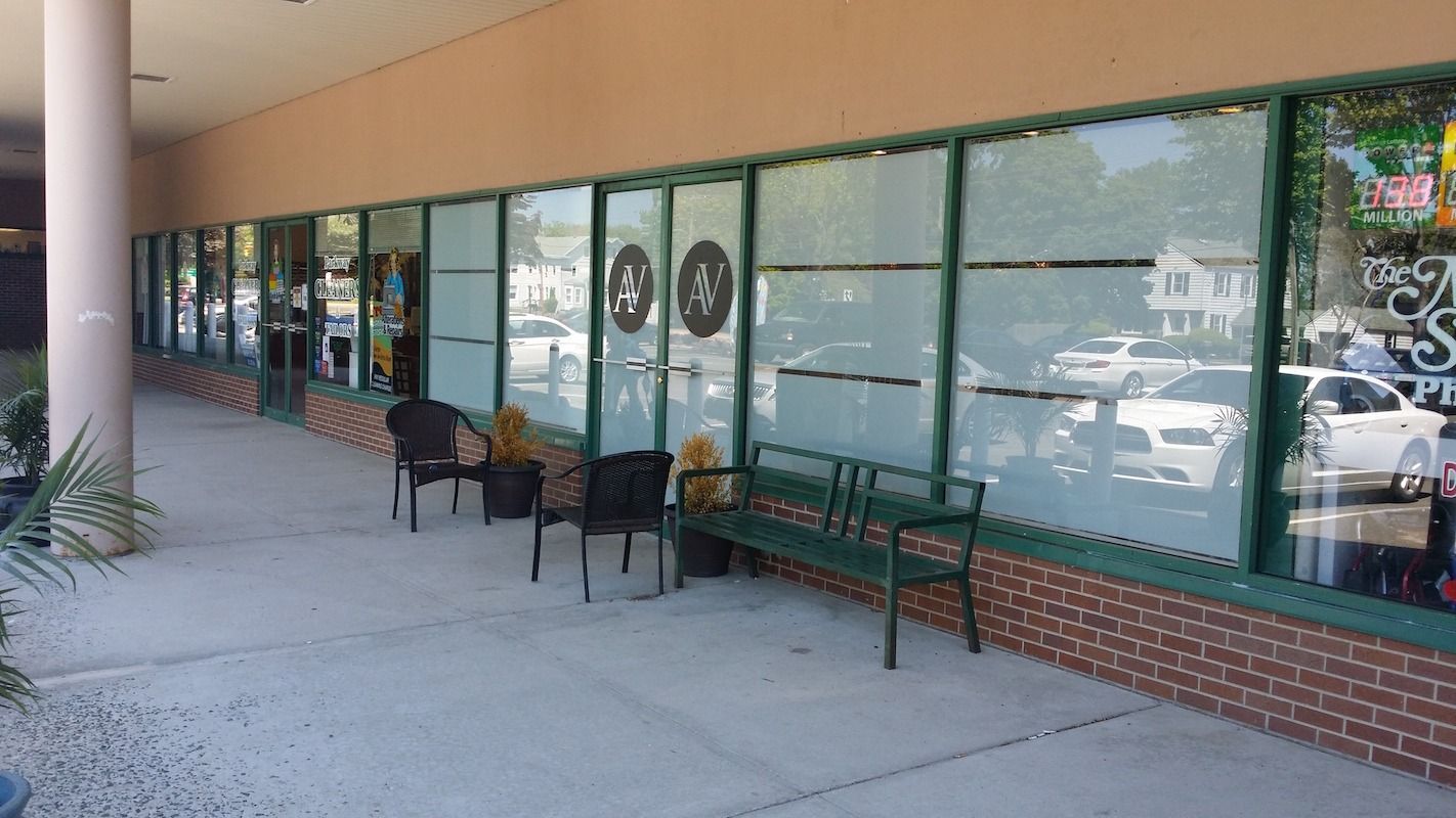 Exterior of a storefront with a curved walkway, seating, and partially frosted windows. Green trim, brick base.