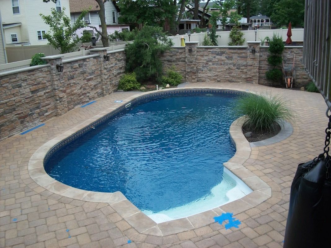 Pool surrounded by stone walls and brick pavers. Blue water, white steps, and greenery.