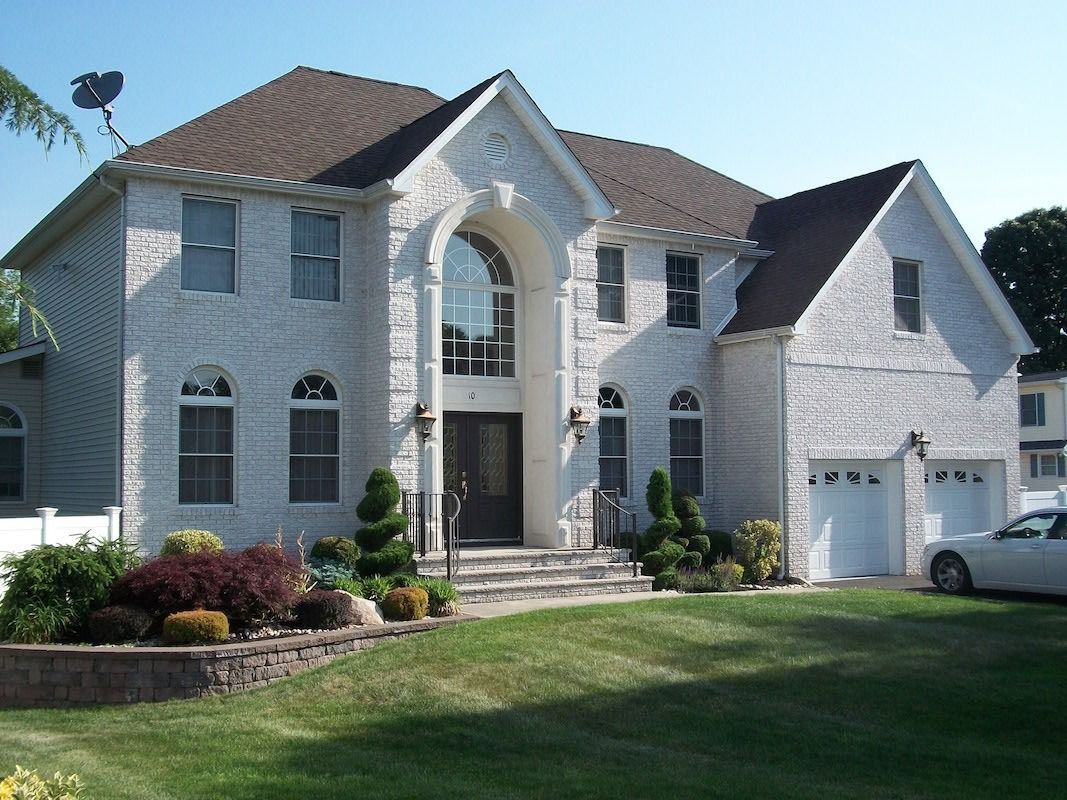 White stucco house with brown roof, two-car garage, and green lawn. Landscaped front yard with bushes.