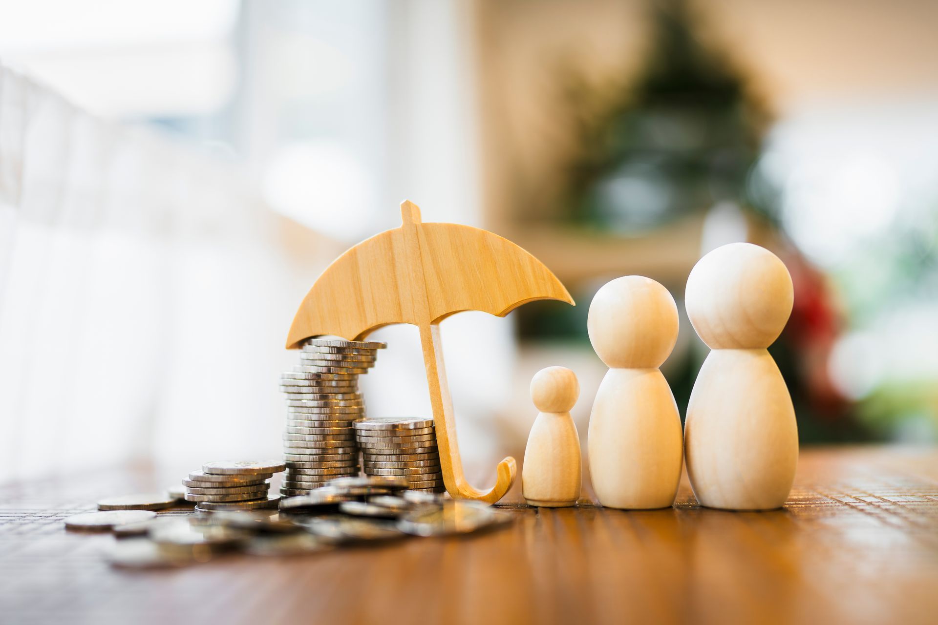 Wooden family figures under a yellow umbrella beside stacked coins on a table