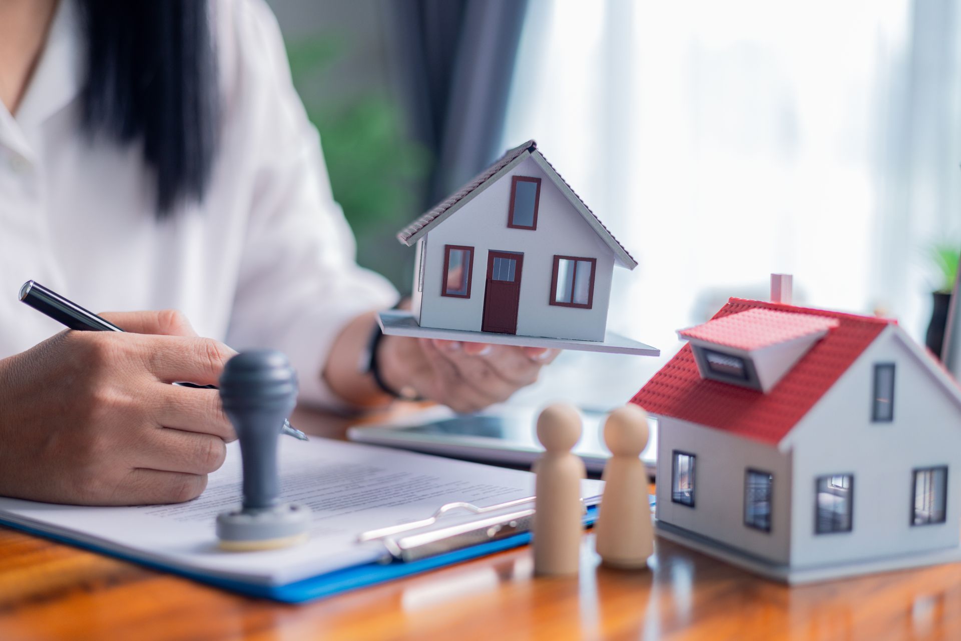 Hand placing a house model beside a notebook, with two small houses on a desk and a stamp-like tool in view