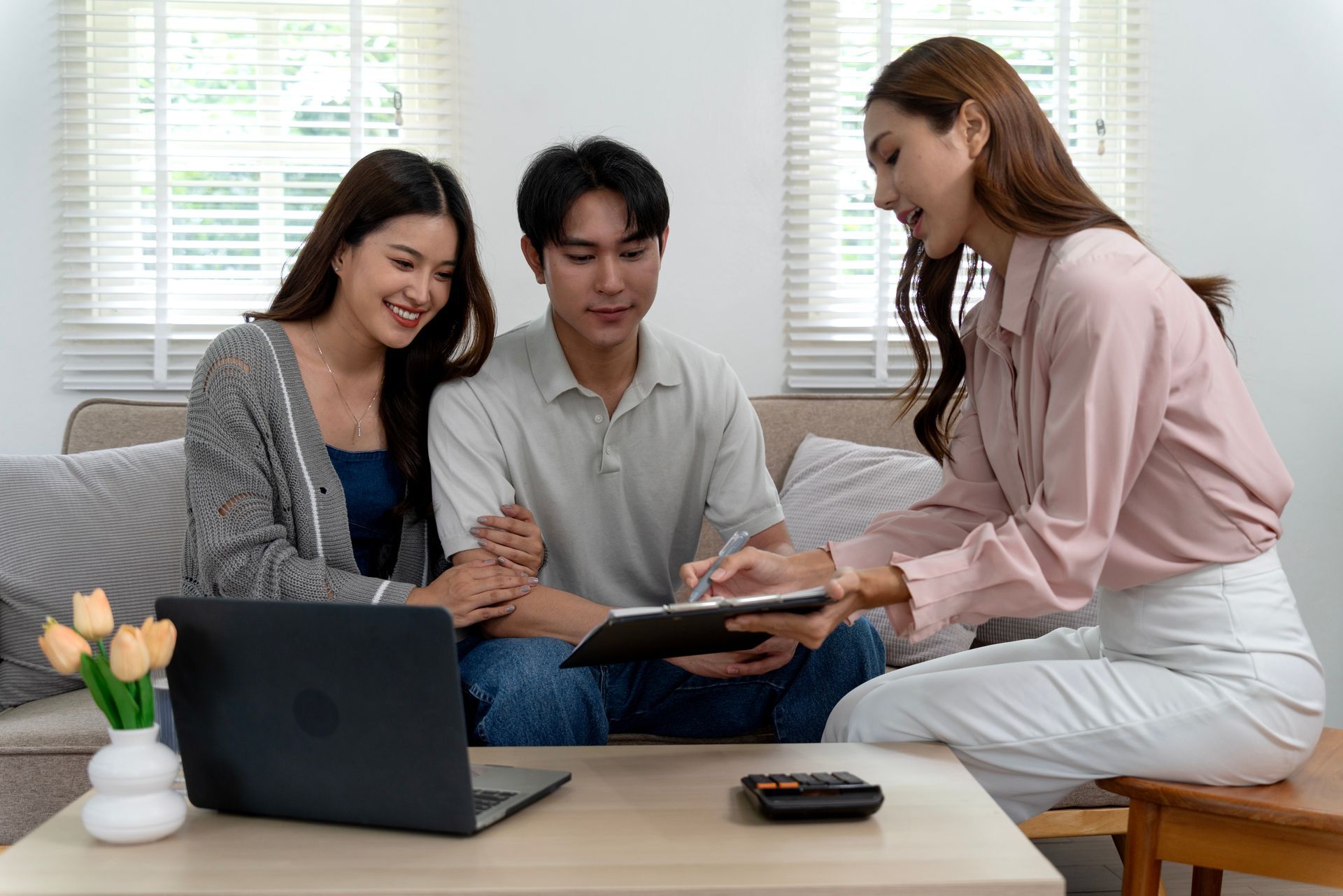 Three people collaborate on a laptop and tablet at a coffee table in a bright living room.