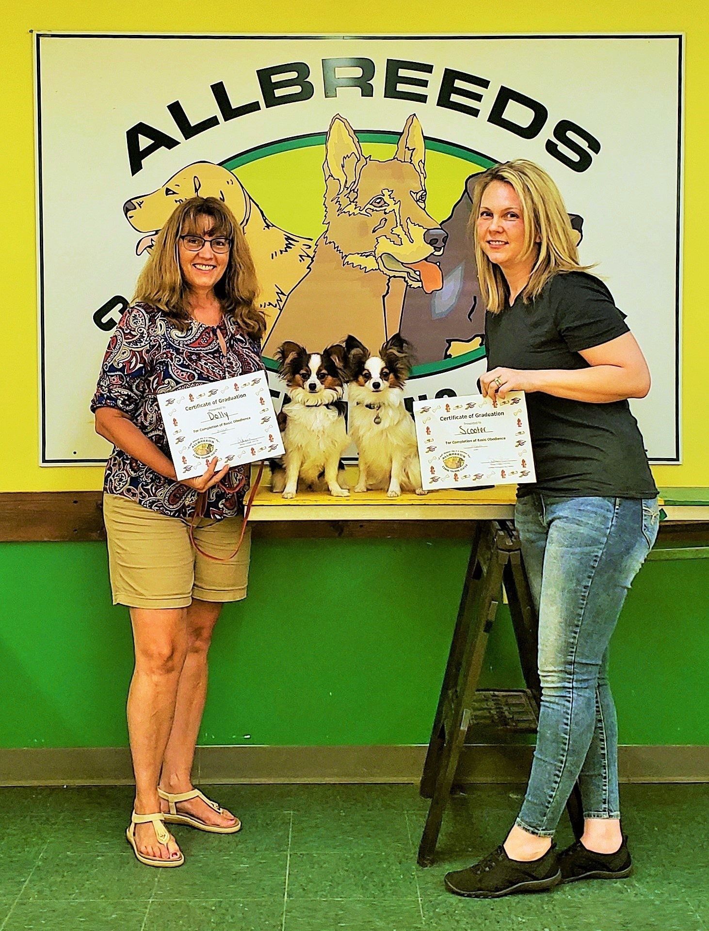Two women with certificates pose with two small dogs in front of an 