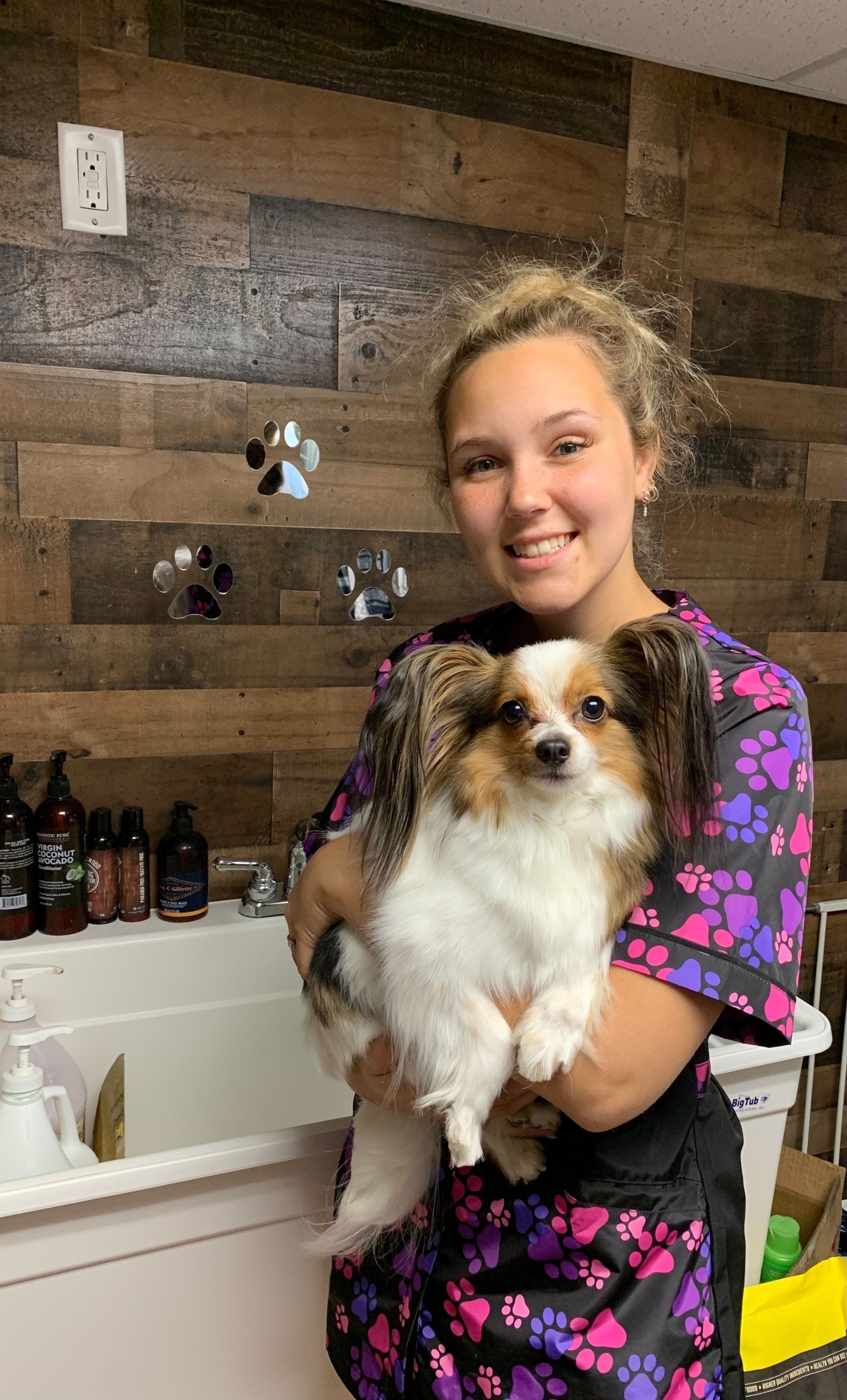 A young woman in a dog grooming apron smiles while holding a small, fluffy, tri-color dog.