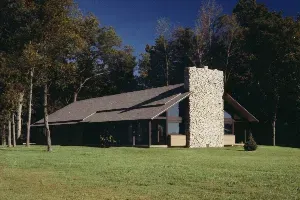 A large house with a large stone chimney in the middle of a lush green field surrounded by trees.