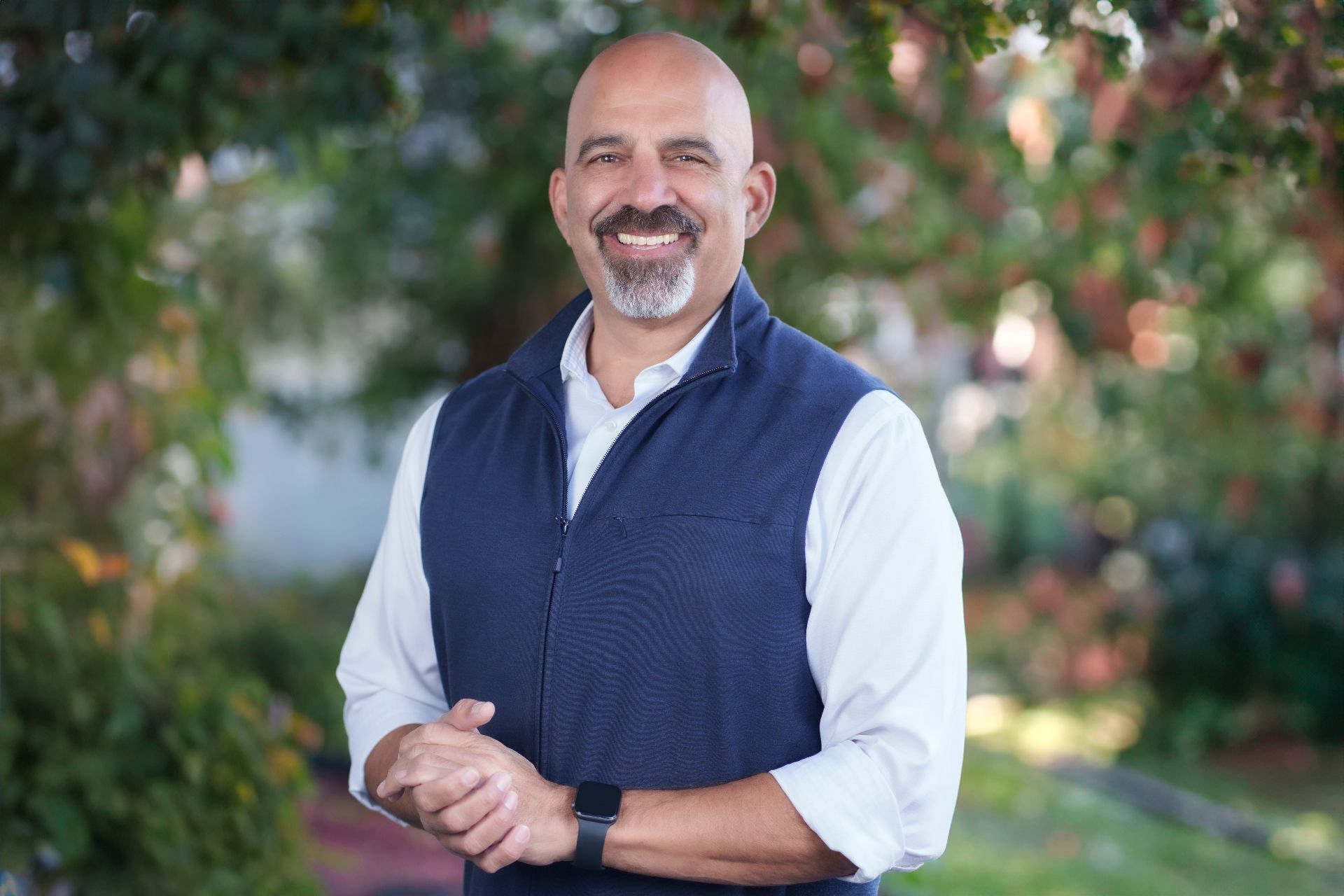 A smiling man with a shaved head and a goatee, wearing a white shirt and dark blue vest, stands outdoors.
