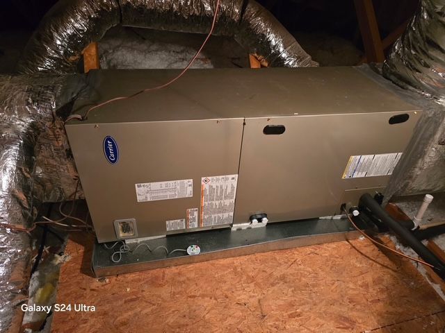 An HVAC unit in an attic, with ductwork and a metal base. The unit is tan and has a Carrier logo.