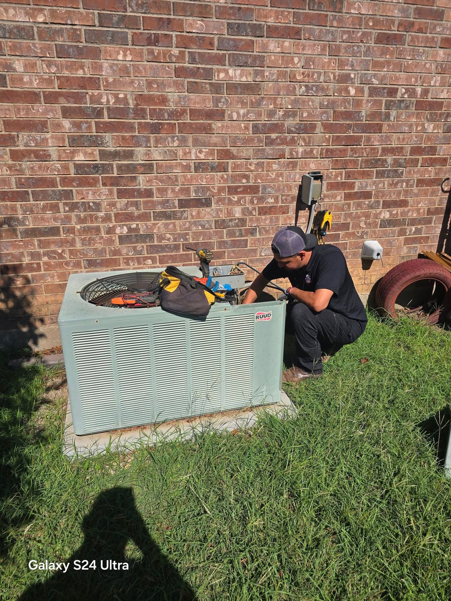 A person repairs an air conditioning unit outdoors. The unit is green, and the background has a brick wall.