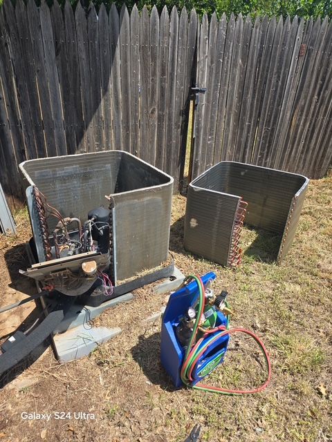 Deconstructed air conditioning unit with torch and tanks in a yard near a wooden fence.