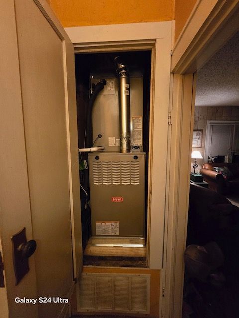 Furnace unit inside a closet, viewed from doorway. Gray metal with silver chimney. Beige walls and door frame.