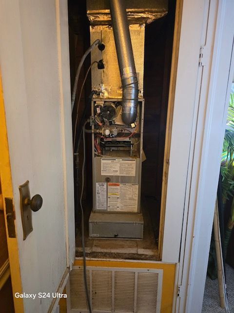 Furnace in a cabinet with open door. Gray and silver components, with vent pipe. Wooden cabinet.